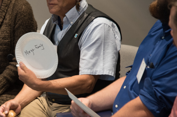 Three people sitting, with the main focus being on one person sharing a thought; and a paper plate saying "Mega-Family"