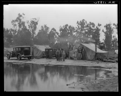 several people stand in front of fabric tents, a truck, scraggly trees, with a huge puddle in the foreground