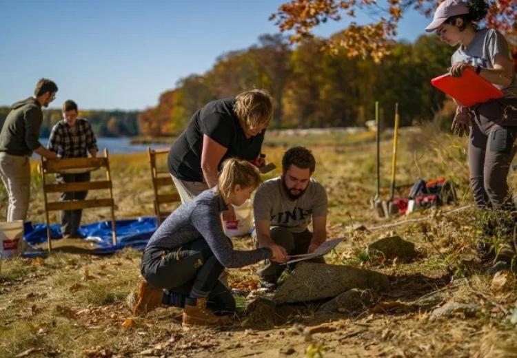 People at work on archaeological dig site, trees and bay in background