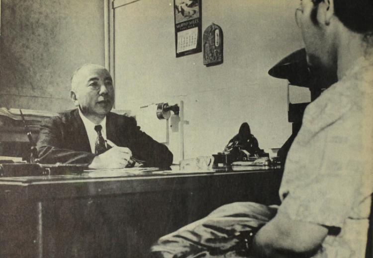 Two men seated, facing each other, one behind desk