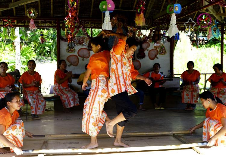 A man and woman are performing the traditional Philippine folk dance Tinikling, which involves a pair of two bamboo poles.