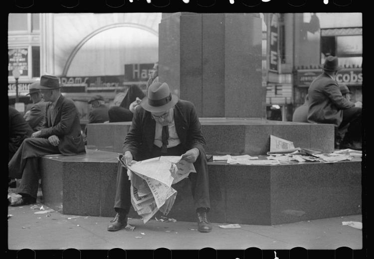 Man sitting on a stone block, reading newspaper. Other men sitting in the background