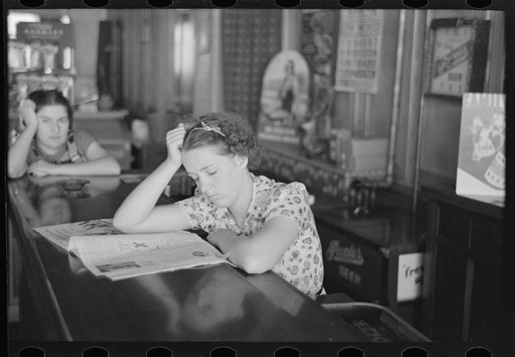 Girl reading newspaper at a counter, resting her head on her hand. Another girl sits in the background.